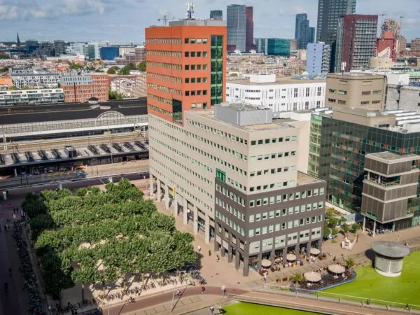Luchtfoto van het Johanna Westerdijkplein in Den Haag met omliggende kantoorgebouwen, terrasjes en het station op de achtergrond.